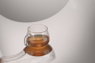 A transparent glass cup with tea on a reflecting surface and a light gray background