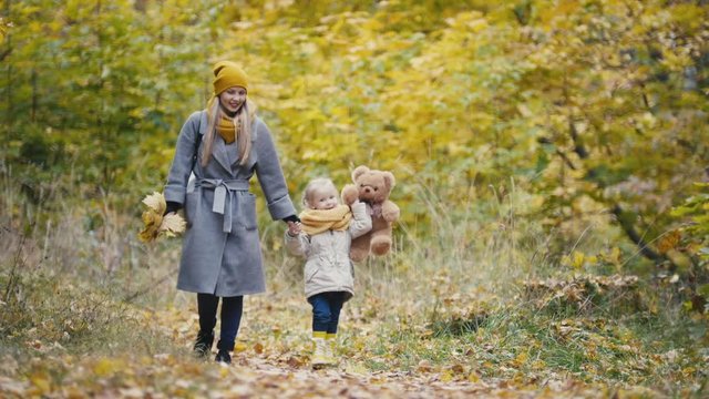 Little daughter with her mommy and Taddy bear walks in autumn park - plays the leaves
