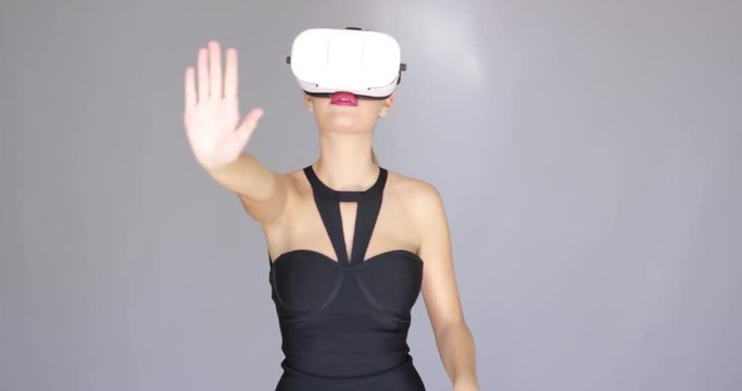 Beautiful sexy blond girl enjoys world in virtual reality glasses. She wearing vr helmet, looking around and gesturing with her hands. Experiencing something exciting. Isolated on gray.