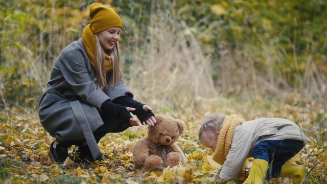 Happy family concept - little daughter with her mommy and Taddy bear plays with yellow leaves in autumn park