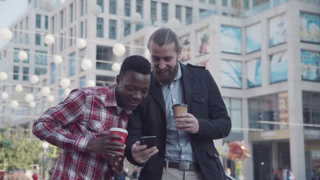 Two different people caucasian and afro american talking to each other till coffee break. One shows to another smartphone picture. Urban background.