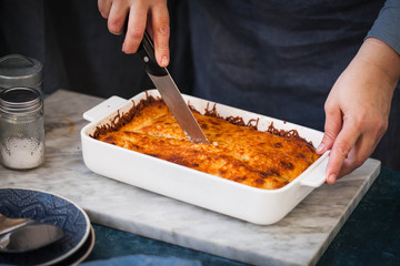 Hot tasty lasagna in ceramic casserole dish. Woman cut with knife hot lasagna. Lasagna after baking. 
