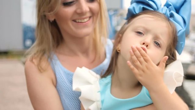little girl three years sitting on the lap of the mother with long blond hair ,which is smiling and looking forward interested and sends a kiss