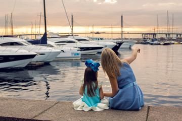 mom shows daughter three years ships in a blue dress and blue big bow sitting on the dock by the...