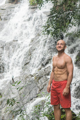 Handsome man with beard wearing red shorts standing and looking up near waterfall. Male tourist enjoying by a water fall in forest