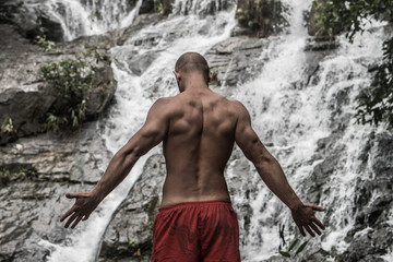 Fototapeta na wymiar Back view of muscular man wearing red shorts standing with raised arms near waterfall. Male tourist enjoying by a water fall in forest