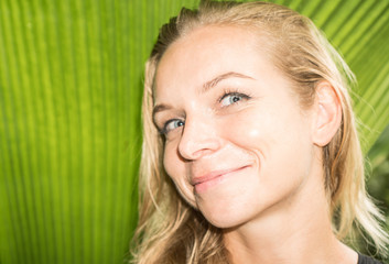 Fototapeta premium Closeup of happy blond woman looking into camera over green tropical leaf background