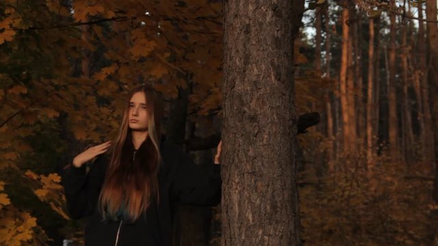A girl in a hoody with her hair loose who holds a tree with her hand takes the hood off and looks around sullenly on the prowl.In a dark forest.