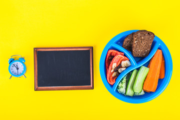 Lunch box with breakfast, board and alarm clock. Time to eat. On a yellow background.