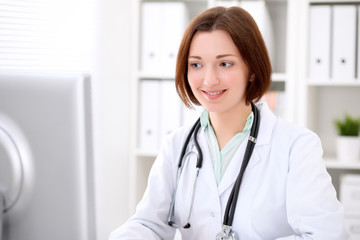 Young brunette female doctor sitting at a desk and working on the computer at the hospital office.  Health care, insurance and help concept. Physician ready to examine patient