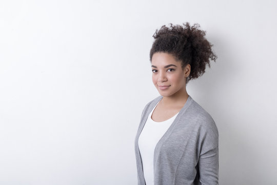 Young African American woman with black curly hair, wearing gray cardigan against white wall 