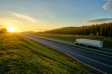 Asphalt highway with a ride white truck between green meadows and larch forest in autumn landscape at sunset.
