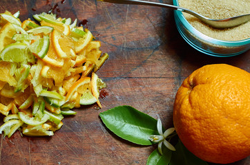 An orange, orange blossom and a pile of sliced oranges, lemons and limes on a wooden board with a bowl of sugar and a spoon.