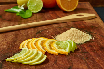 An orange, a half orange, a half lime and a lemon on a wooden board with a wooden spoon, orange slices, lime slices, lemon slices and a pile of brown sugar.