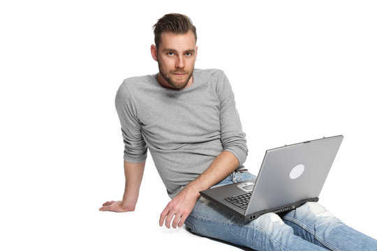 A man in his 20s sitting down on the floor with his laptop, wearing jeans and a grey shirt. White background.