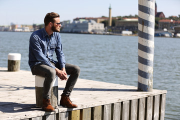 A depressed man sitting down outside on the dock with the ocean behind him, wearing jeans and a...