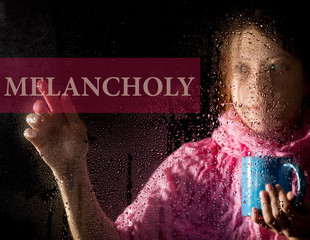 melancholy written on virtual screen. young woman melancholy and sad at the window in the rain, she holding a cup of hot coffee or tea