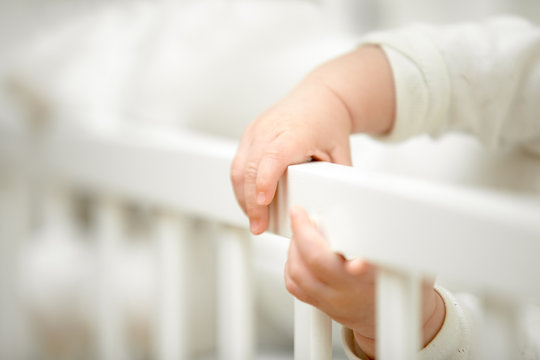 Infant Tiny Hands In The Crib Holding Onto The Side Of The Bed, Have Fun Grabbing It. Closeup, Baby Development Concept Photo