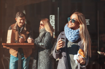 Beautiful young blonde woman in sunglasses holding coffee cup and croissant and looking at sun with smile while standing outdoor. The weather is great today to walk with friends!