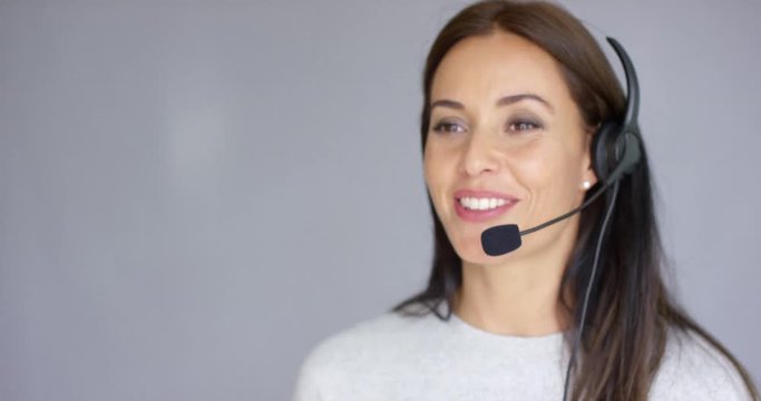 Adorable and beautiful female call center agent speaking with someone on headset. She has smile on her face. Isolated on gray.