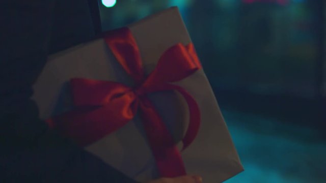 CU FOLLOW Young Caucasian male carries a present white gift box with red ribbon bow, urban background. 4K UHD