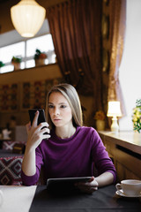 Woman looks thoughtfully in smartphone holding tablet. Vertical indoors shot. 