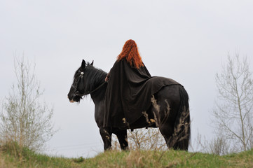 Fototapeta premium Brave woman with red hair in black cloak on friesian horse