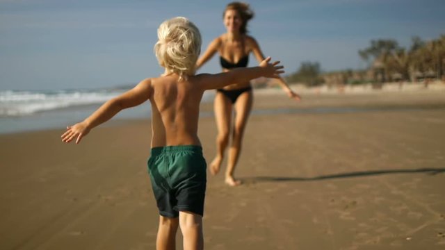 A cute boy runs a long the beach to her mothers open arm's as the camera follows him. Slow motion