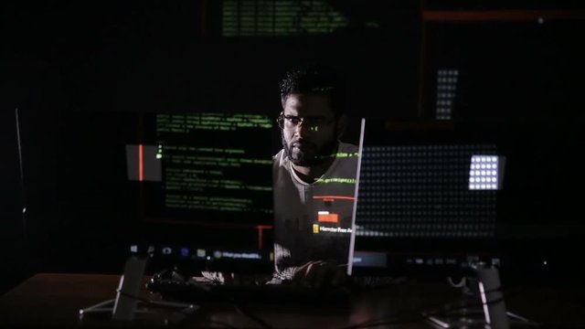Male hacker hacks computer in dark. Computer code reflecting on his face. HD.