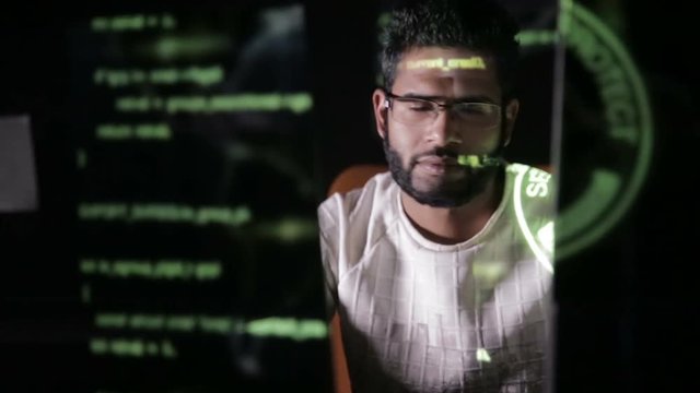 Male hacker hacks computer in dark. Computer code reflecting on his face. HD.