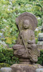Fototapeta na wymiar Tokyo, Japan - September 26, 2016: Closeup of stone statue of Bodhisattva sitting on lotus pedestal in garden at Senso-ji Buddhist Temple. Large halo. Holds staff and sphere. Green foliage. 
