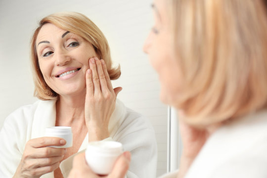 Senior woman applying cosmetic cream on face near mirror