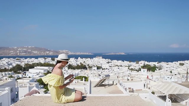Beautiful woman with smart phone outdoors in old streets an Mykonos. Girl at street of typical greek traditional village with white walls and colorful doors on Mykonos Island, in Greece, Europe