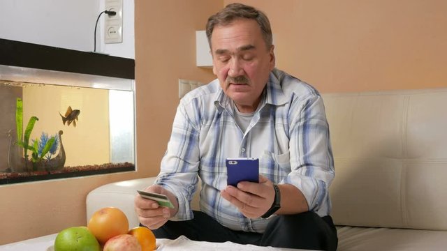 Senior man with a mustache makes a purchase from the online store credit card. He was sitting at home on the couch and enters the card information to phone