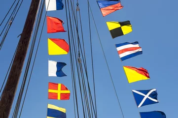 Abwaschbare Fototapete Segeln Colorful nautical sailing flags flying in the wind from the lines of a sailboat mast backlit in bright blue sky by the sun