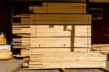 Stacks of thin planks stored outside a building partly shaded by the roof.
