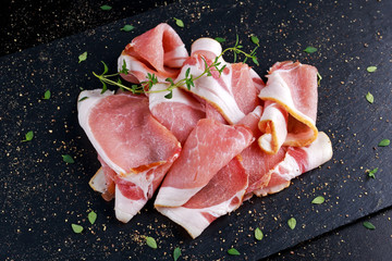Raw smoked bacon slices on black stone board which herbs