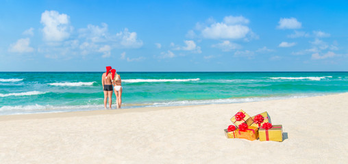 Couple in Christmas Santa hats at beach with holiday gifts