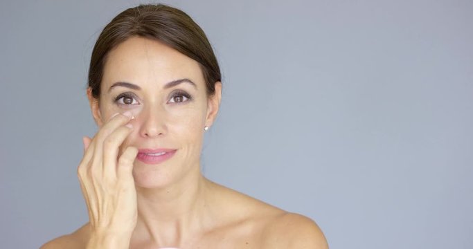 Beautiful young woman applying face cream to her cheek bone in a beauty  skincare or cosmetics concept  on grey with copy space