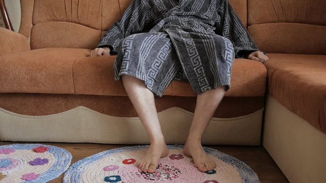 The white man in the bathrobe sitting on the sofa and stamping his feet. Unrecognizable person.