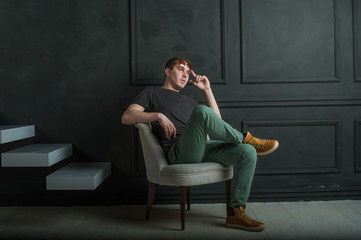 studio photography young brutal guy. man in sunglasses, T-shirt, jeans and high boots on a background of black wall, He is sitting in a white armchair throne.