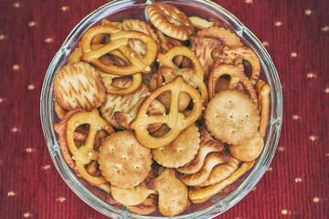 Mix of salty snacks (crackers and pretzels) in glass bowl on red background