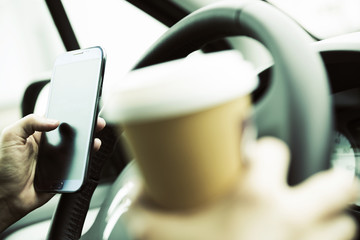 Geschäftsfrau mit einem Becher Kaffee in der Hand bedient ihr Smartphone am Lenkrad im Auto