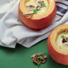 Two servings of pumpkin soup in bowls made of mini-pumpkins, on green background, garnished with marjoram, pumpkin seeds and red pepper, ready to be eaten – seasonal food concept 