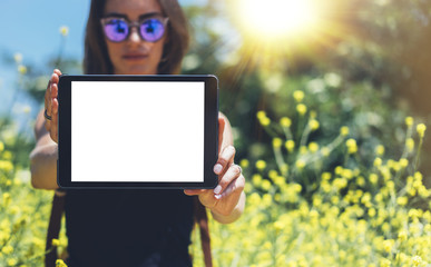 Hipster holding in hands tablet computer. Girl traveler with sunglasses using gadget on sun flare and yellow flowers background, mockup of blank empty screen copy space for text, female photographer