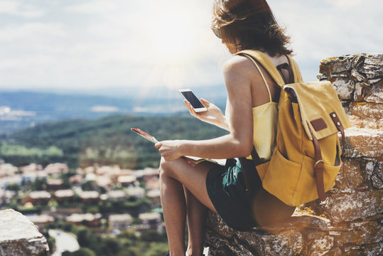 Hipster young girl with bright backpack looking at phone and map. Said view tourist traveler on background mountain, blue sea, sun flare. Mockup for text message. Female hands using smartphone