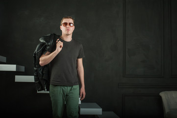 studio photography young brutal guy. man in sunglasses, T-shirt, jeans keeps black leather jacket, slung over his shoulder. black background painted walls and white stairs