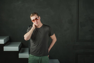 studio photography young brutal guy. man in sunglasses, T-shirt, jeans keeps black leather jacket, slung over his shoulder. black background painted walls and white stairs