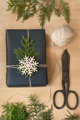 Christmas gift box wrapped in black paper and twine around branch cypress on wooden surface.