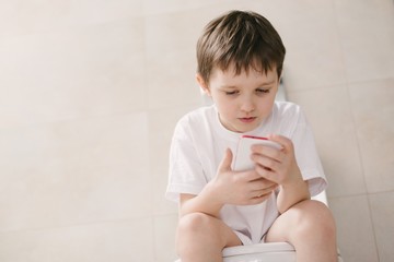 Boy playing with smartphone while sitting on toilet. - obrazy, fototapety, plakaty